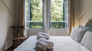 A bedroom with a bed, two windows, a chair, and a view of trees. - Casa da Tia Luísa (Furnas)