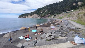 A beach with thatched umbrellas, people sunbathing, and a rocky shoreline. - Casa da Tia Luísa (Furnas)