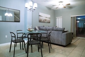 Un coin repas avec une table en verre et quatre chaises, un canapé gris, un lustre et un ventilateur de plafond.
