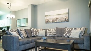 Una sala moderna con un sofá seccional gris, almohadones estampados, una mesa de centro oscura y un cuadro grande de paisaje en la pared.