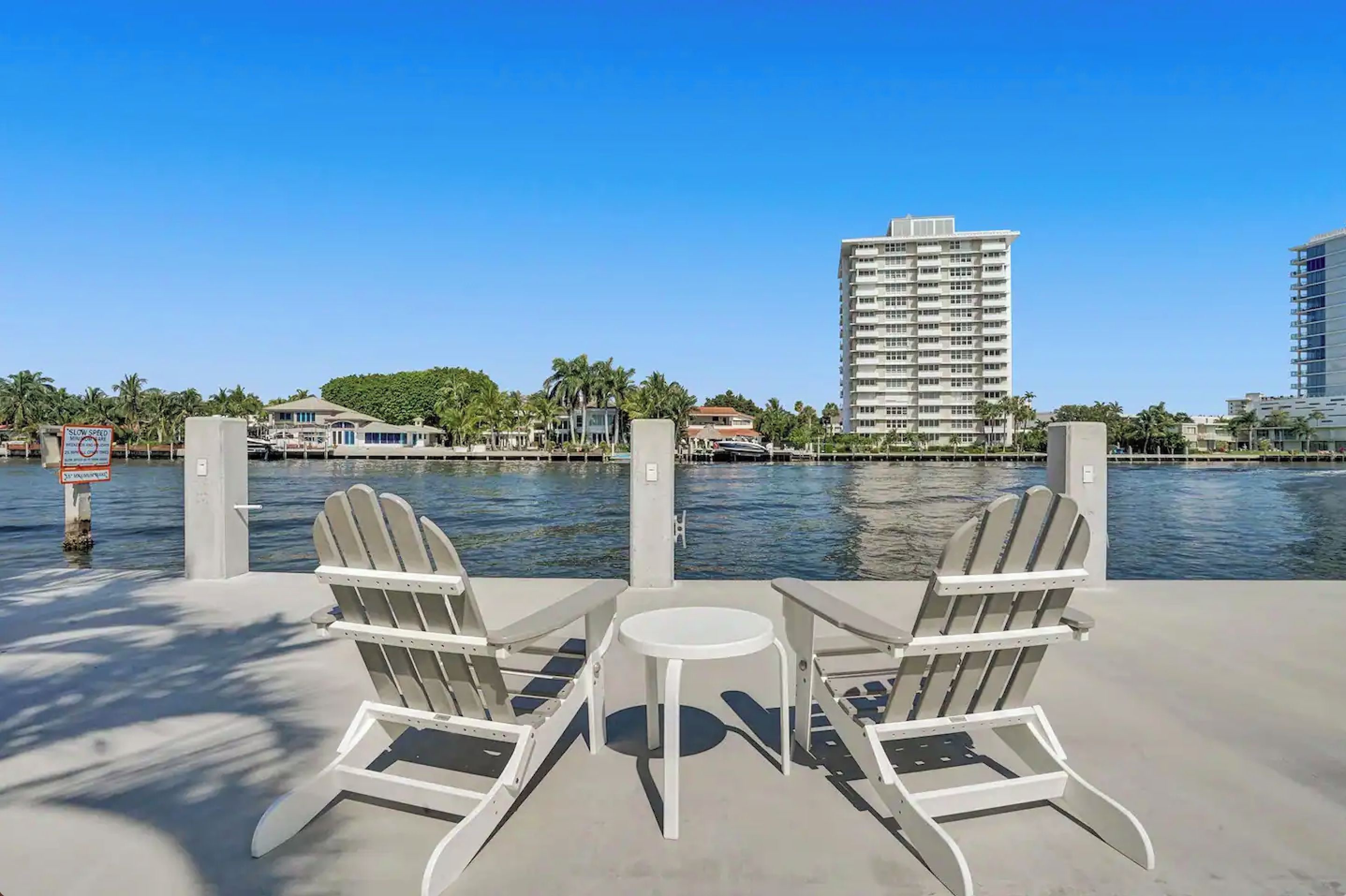 A waterfront patio with Adirondack chairs and a small table, overlooking a body of water and a cityscape with tall buildings.