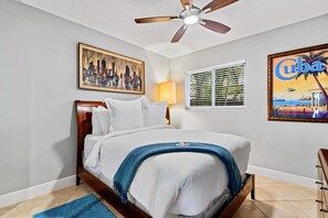 A bedroom with a bed, a ceiling fan, a cityscape painting, a lamp, a window with blinds, and a poster of Cuba. - Wilton Manors 2BR Charm! Walk to the Drive (Wilton Manors)