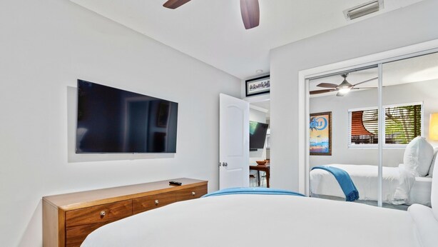 A modern hotel room with a large bed, a wooden dresser, a flat-screen TV mounted on the wall, and a sliding glass door leading to a balcony with a view of the city. - Wilton Manors 2BR Charm! Walk to the Drive (Wilton Manors)