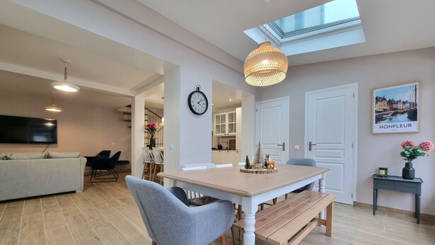 Een moderne eetruimte met een houten tafel, grijze stoelen en een hanglamp. Een klok hangt aan de muur, en er is een lijst met een stadsgezicht.