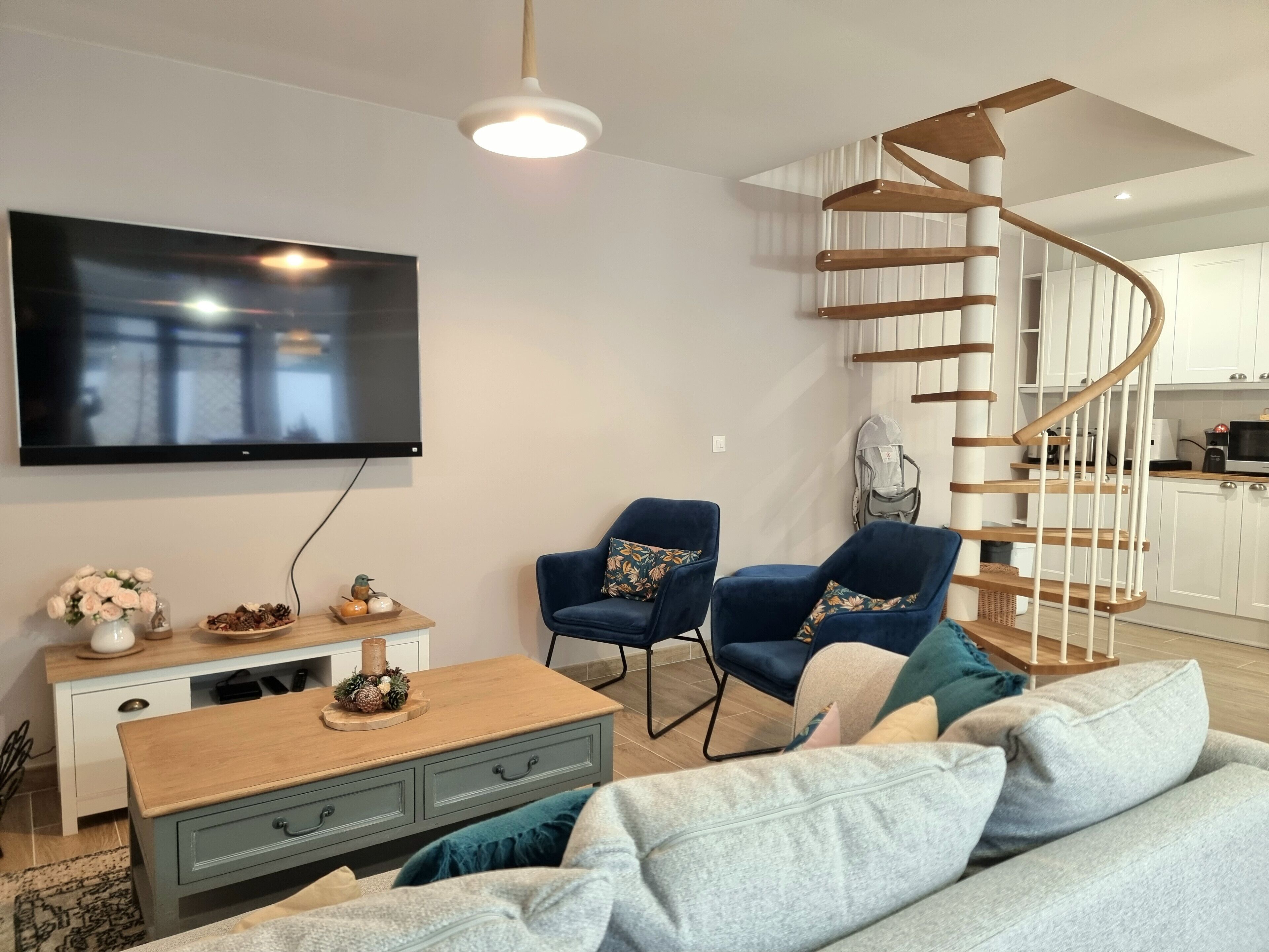 A modern living room with a flat-screen TV, two blue armchairs, a grey sofa, a wooden coffee table, and a spiral staircase leading to a kitchen area.