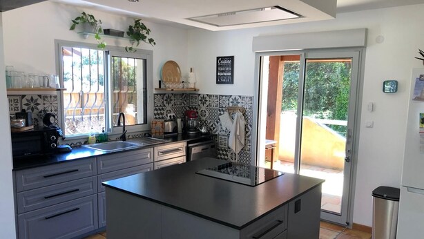Una cocina moderna con una isla central, un fregadero y un refrigerador. Hay plantas en macetas en el alféizar de la ventana y una pizarra con escritura en la pared.