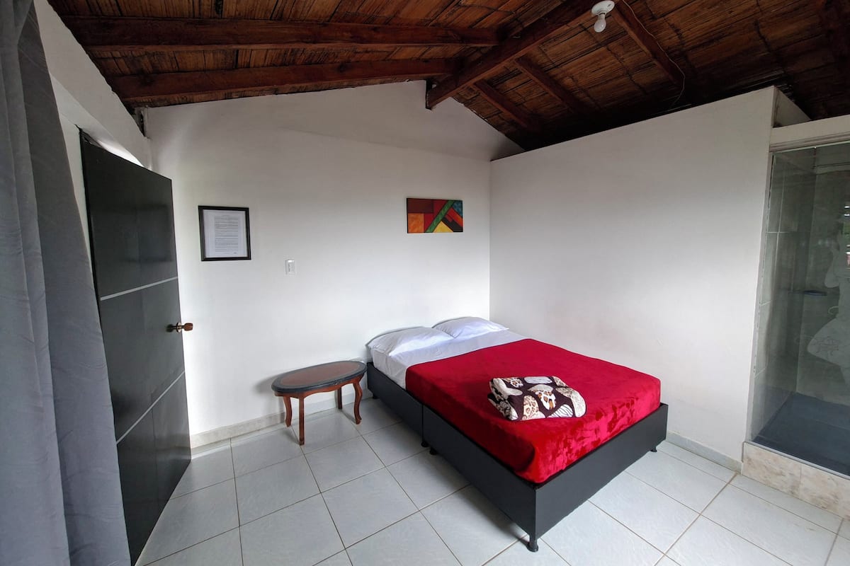 A bedroom with a wooden ceiling, a bed with a red bedspread, a small table, and a framed picture on the wall.