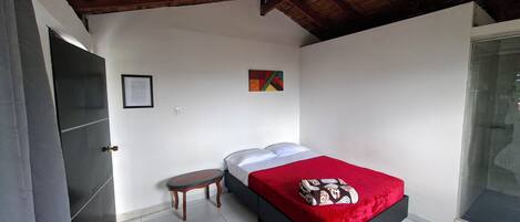 Un dormitorio con techo de madera, una cama con colcha roja, una mesita y un cuadro enmarcado en la pared.