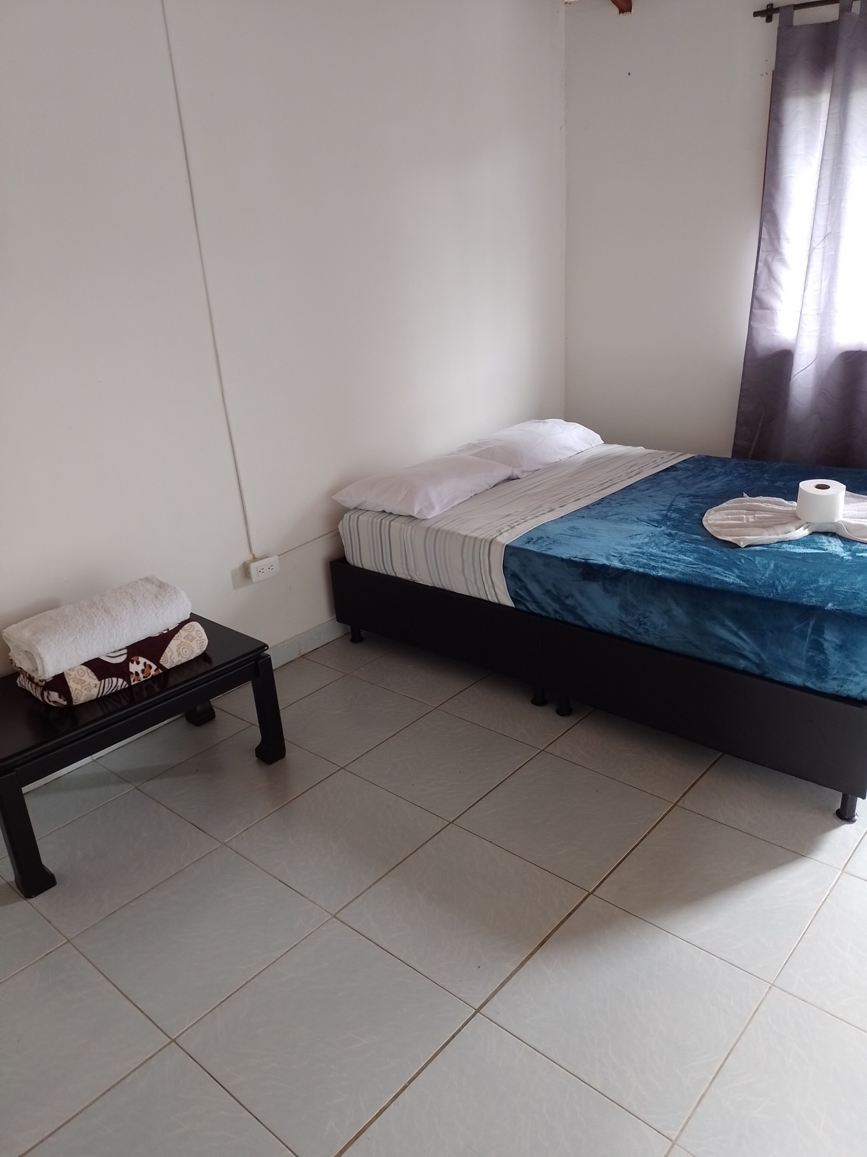 Un dormitorio con una cama, una mesita con una toalla y una cortina.