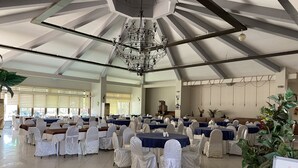 Un salón amplio con filas de mesas y sillas, una araña y una pizarra con texto.
