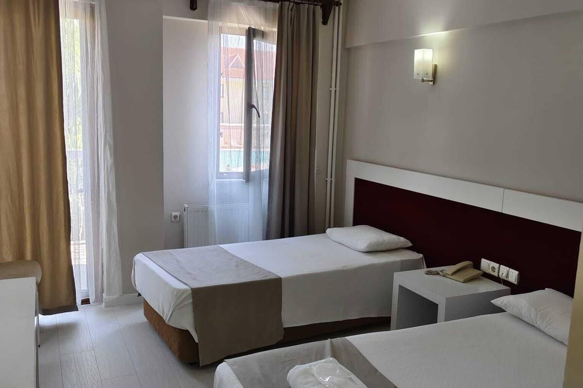 A hotel room with two beds, a bedside table with a phone, a window with curtains, and a wall-mounted light.