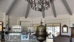Un vestíbulo amplio con techo alto, una lámpara de araña metálica decorativa, un mostrador de recepción con separador de vidrio, un gran jarro metálico ornamentado y un sofá color beige.