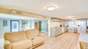 Un salón amplio con un sofá color beige, una cocina con electrodomésticos blancos y un suelo de madera claro.