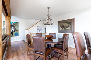A dining area with wooden chairs, a round table, and a chandelier. There is a painting on the wall and a staircase in the background. - Saddle Ridge V2 (3-Bedroom Condo) with Mountain view by Moonlight Basin Lodging (Big Sky)