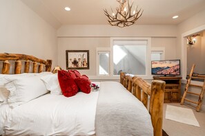 A bedroom with a large bed, a wooden headboard, a chandelier, a television, and a framed picture on the wall. - Saddle Ridge V2 (3-Bedroom Condo) with Mountain view by Moonlight Basin Lodging (Big Sky)