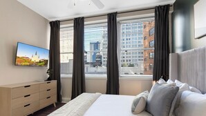 A modern hotel room with a large bed, a TV on the wall, and a city view through the windows.
