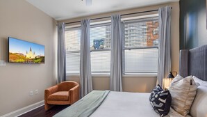 A hotel room with a bed, a chair, a painting, and large windows with blinds.