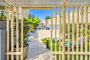 A covered outdoor area with a pathway leading to a seating area, surrounded by greenery and a view of buildings.