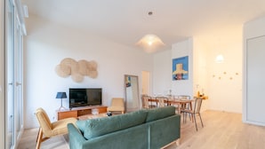A modern living room with a green sofa, wooden flooring, a TV, and a dining area with chairs and a table.
