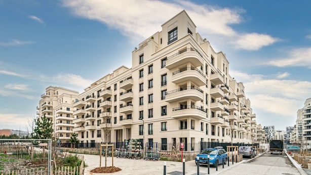 A multi-story residential building with balconies, a parking area, and a clear sky.