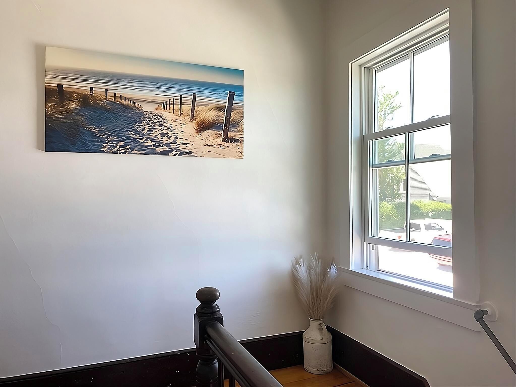 Una escalera con barandilla, un cuadro de una escena de playa y una ventana con vista a una casa y árboles.
