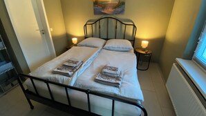 Un lit bien fait, avec deux oreillers et une couverture pliée, une table de chevet avec une lampe, un tableau encadré au mur et un radiateur.