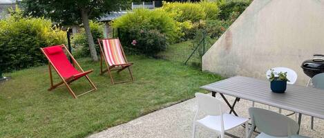 A patio with a table and chairs, two striped lounge chairs, and a garden with bushes and a tree.