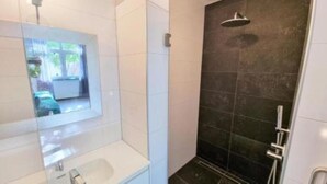 Une salle de bain moderne avec une douche à l’italienne en verre, un lavabo blanc et un mur de douche recouvert de carreaux foncés.