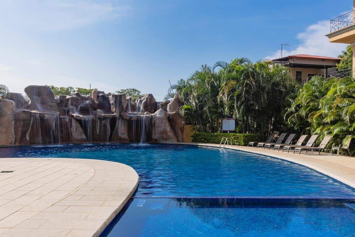 A swimming pool with a waterfall feature, surrounded by palm trees and lounge chairs.