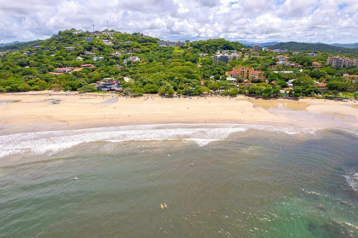 A coastal landscape with a sandy beach, a clear view of the sea, and a hill with buildings and greenery in the background.