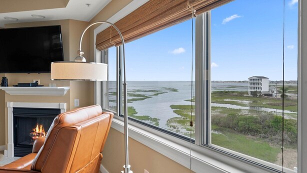 A room with a leather chair, a fireplace, a TV, and a large window with a view of a river and buildings. - 🏖️Serene Bayfront Bliss - Pristine 3 Bed/3Bath condo🏖️ (Ocean City)