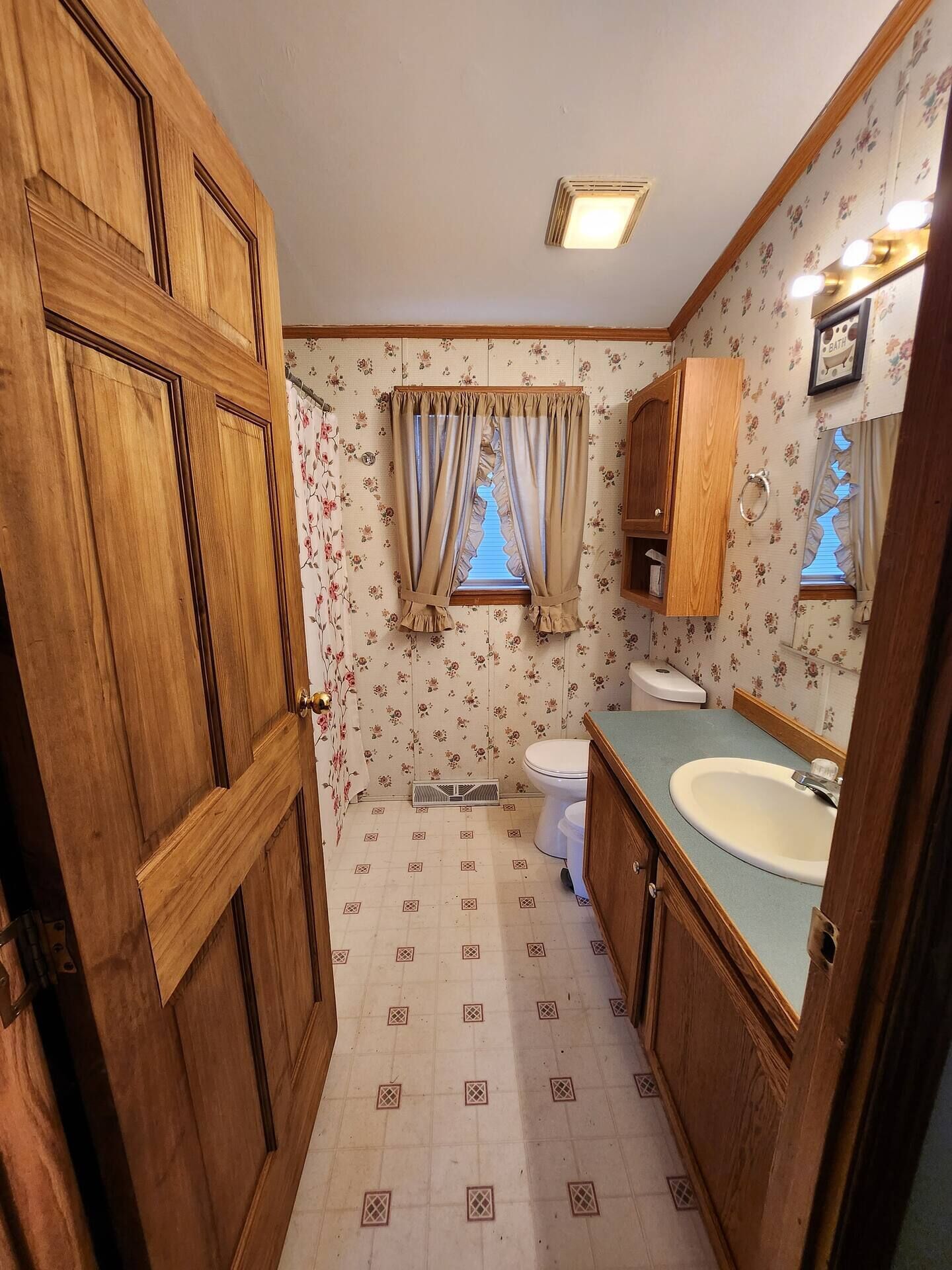 Un baño con muebles de madera, inodoro, lavabo y ventana con cortinas.