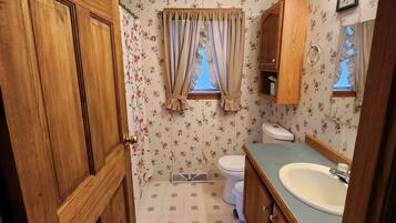 Un baño con muebles de madera, inodoro, lavabo y ventana con cortinas.