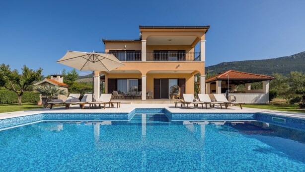 Une maison à deux étages avec une piscine à l’avant, entourée de chaises longues et de parasols.