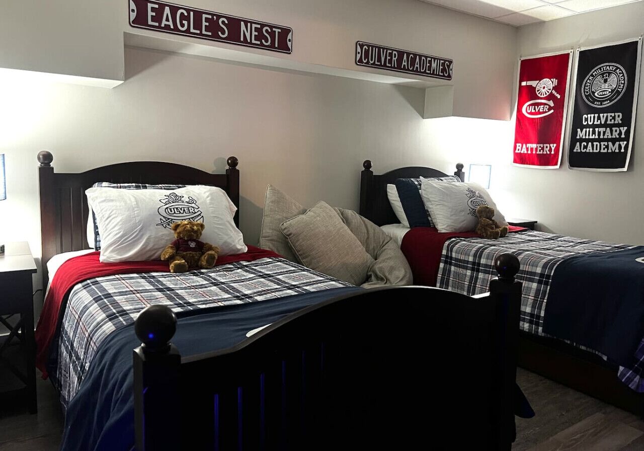 A hotel room with two beds, each with a teddy bear, plaid bedding, and a sign above indicating 'Eagle's Nest' and 'Culver Academy'.
