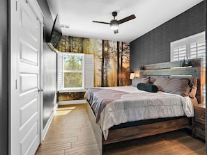 A bedroom with a bed, a ceiling fan, a window, and a wall with a forest mural.