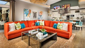 A modern living room with a large orange sectional sofa, a glass coffee table, and artwork on the walls. - Upscale Furnished Resort Home (Rio Verde)