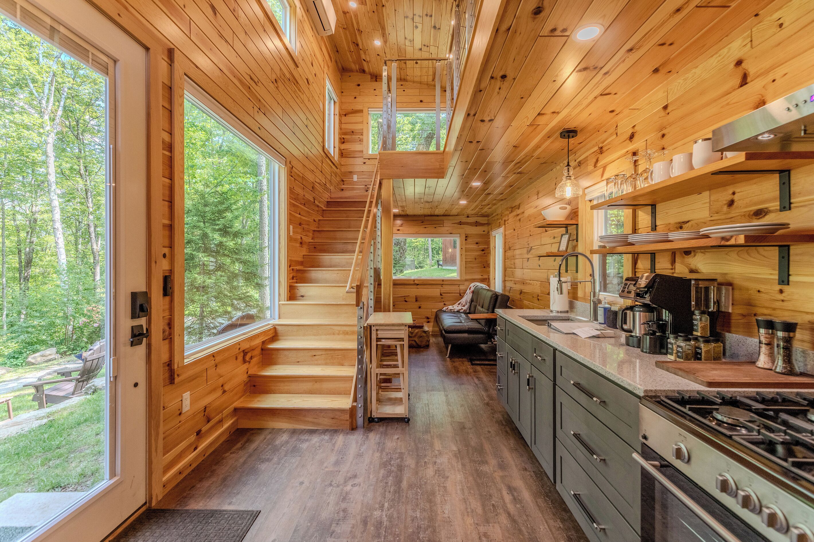 A cabin interior with a wooden staircase, a kitchen with modern appliances, and large windows offering a forest view.