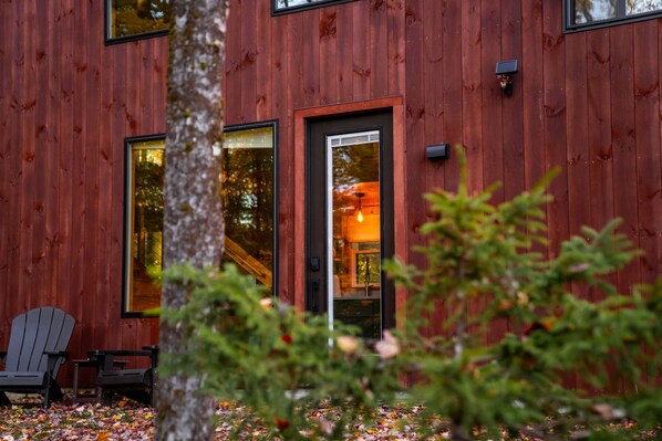 A wooden cabin with large windows, a door, and a small porch with Adirondack chairs. - Luxury Cabin #3 Hot Tub, Kayaks, Paddle Boards, Private Fire Pit (Cavendish)