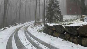 Uma estrada coberta de neve serpenteia por uma floresta, com um muro de pedra e um edifício vermelho ao fundo.