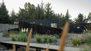 Una cabaña moderna con terraza de madera, rodeada de un paisaje natural con árboles y plantas.