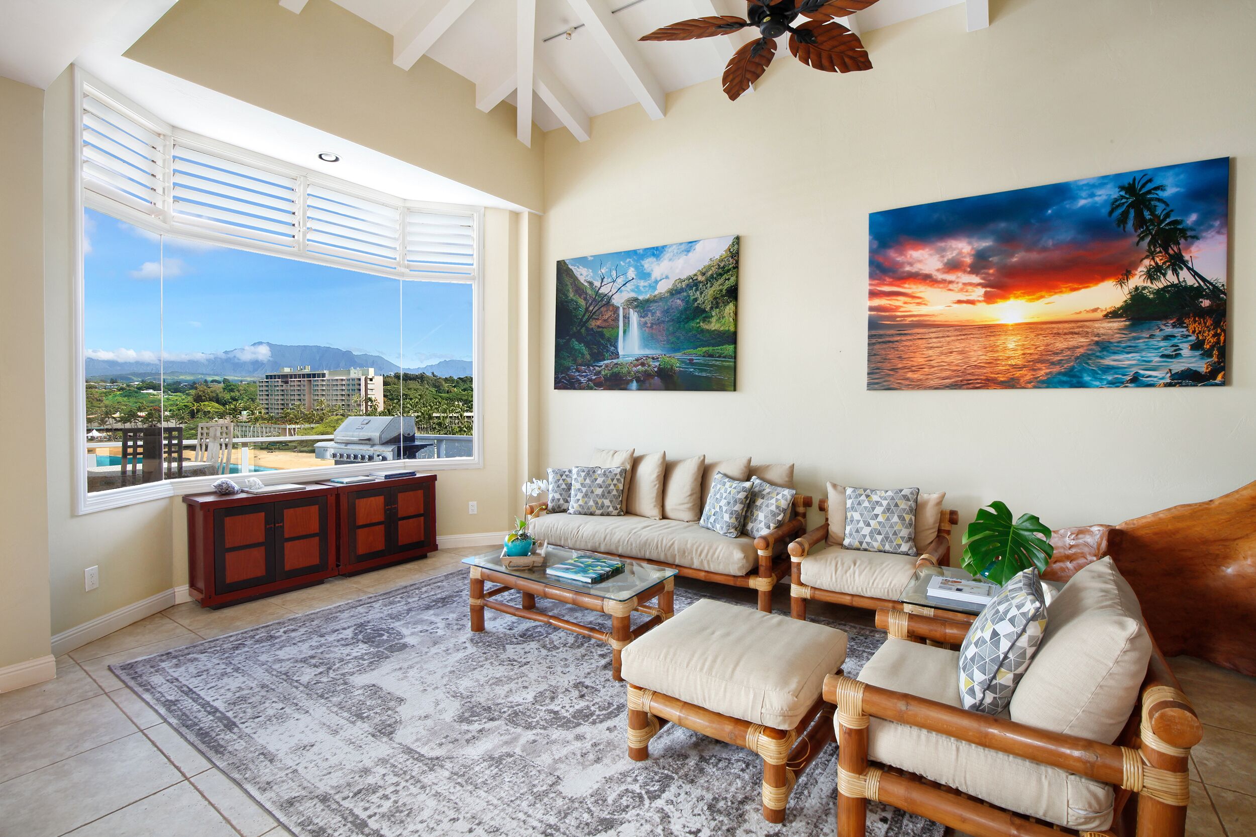 A spacious living room with a large window offering a view of a mountainous landscape, a sofa set, and two paintings on the wall.