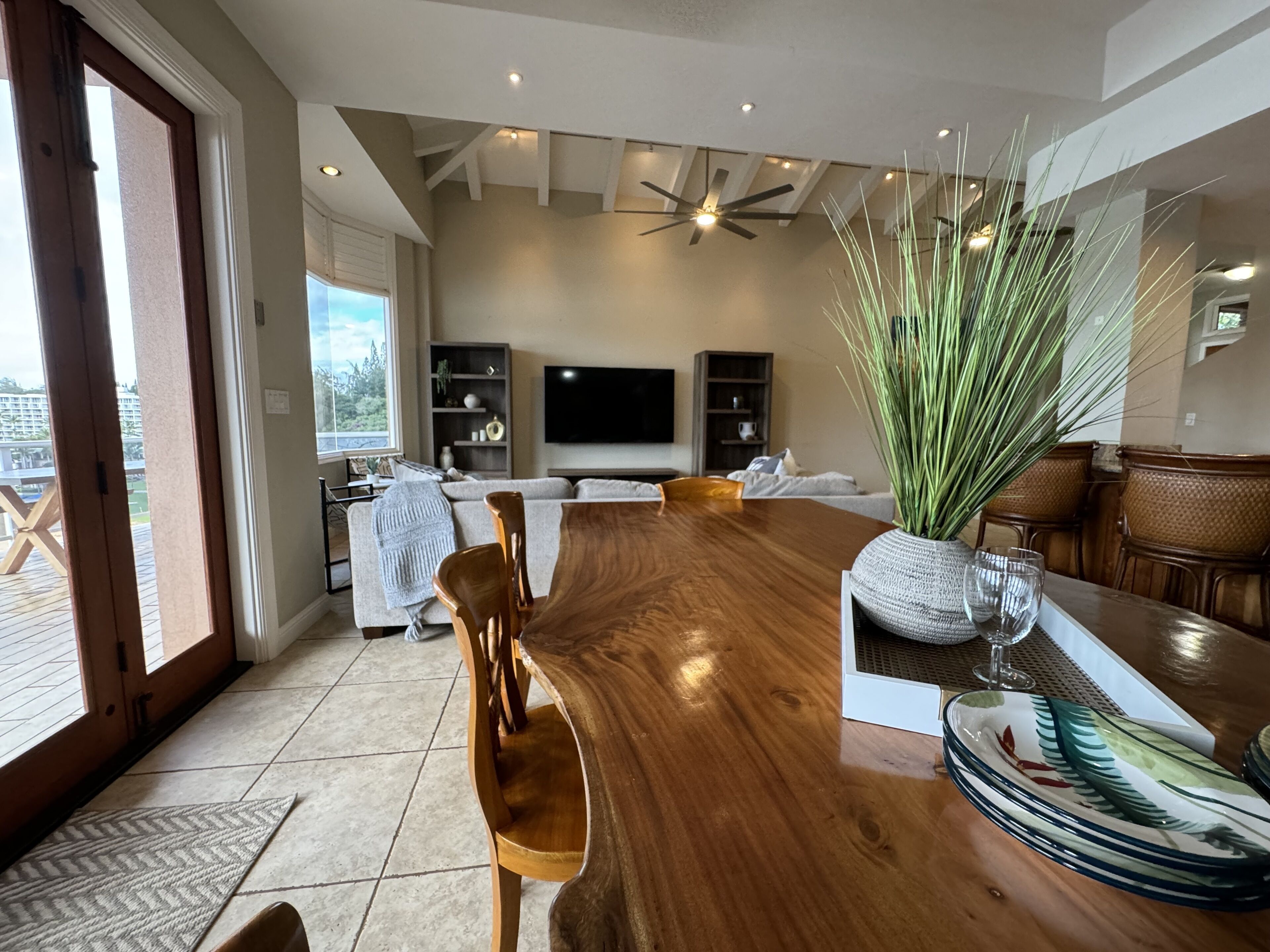 A spacious living room with a wooden dining table, comfortable seating, a large window, and a ceiling fan.