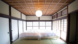 Perinteinen japanilaistyylinen huone, jossa on tatamimatto, matala puinen sänky, liukuovet ja suuret ikkunat.