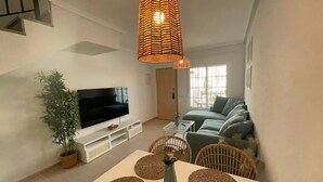 Ein modernes Wohnzimmer mit einem Flachbildfernseher, einem Esstisch mit gewobenen Tischsets, Korbstühlen, einer großen gewobenen Pendelleuchte und einer Topfpflanze.