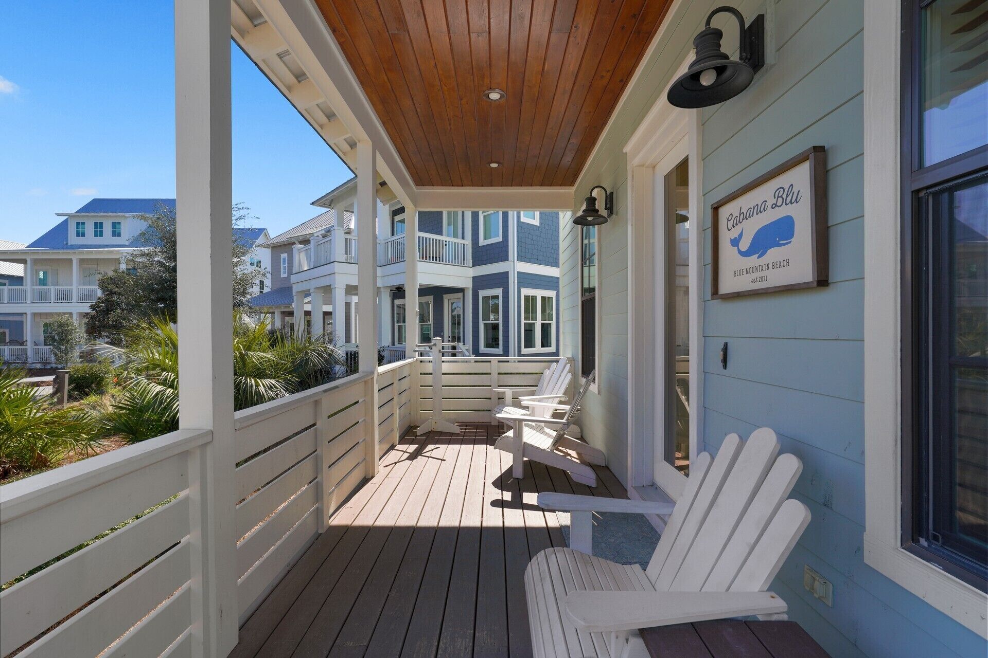 A covered porch with white Adirondack chairs, a wooden ceiling, and a sign reading "Rebecca Blu Blue Mountain Beach."