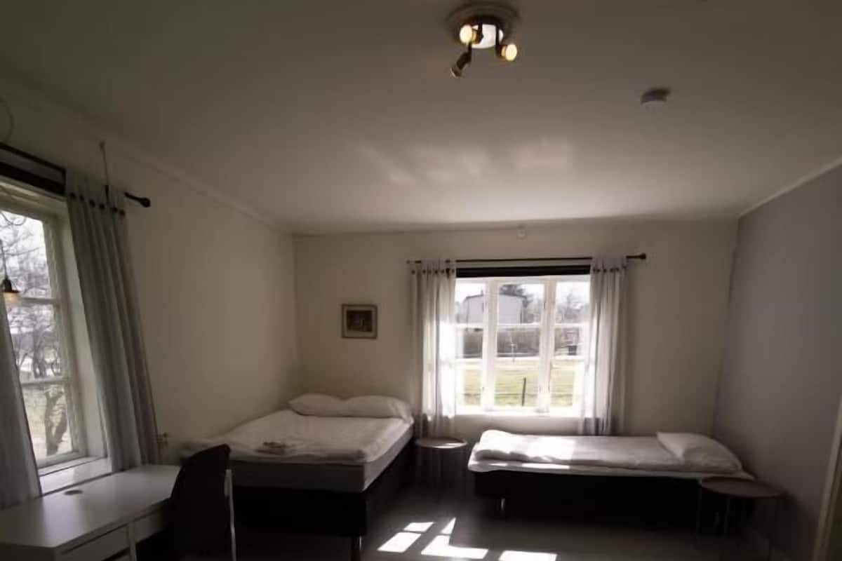 Ein Zimmer mit zwei Einzelbetten, einem Schreibtisch und einem Stuhl. Es gibt ein Fenster mit Vorhängen, eine Deckenleuchte und ein Bild an der Wand.