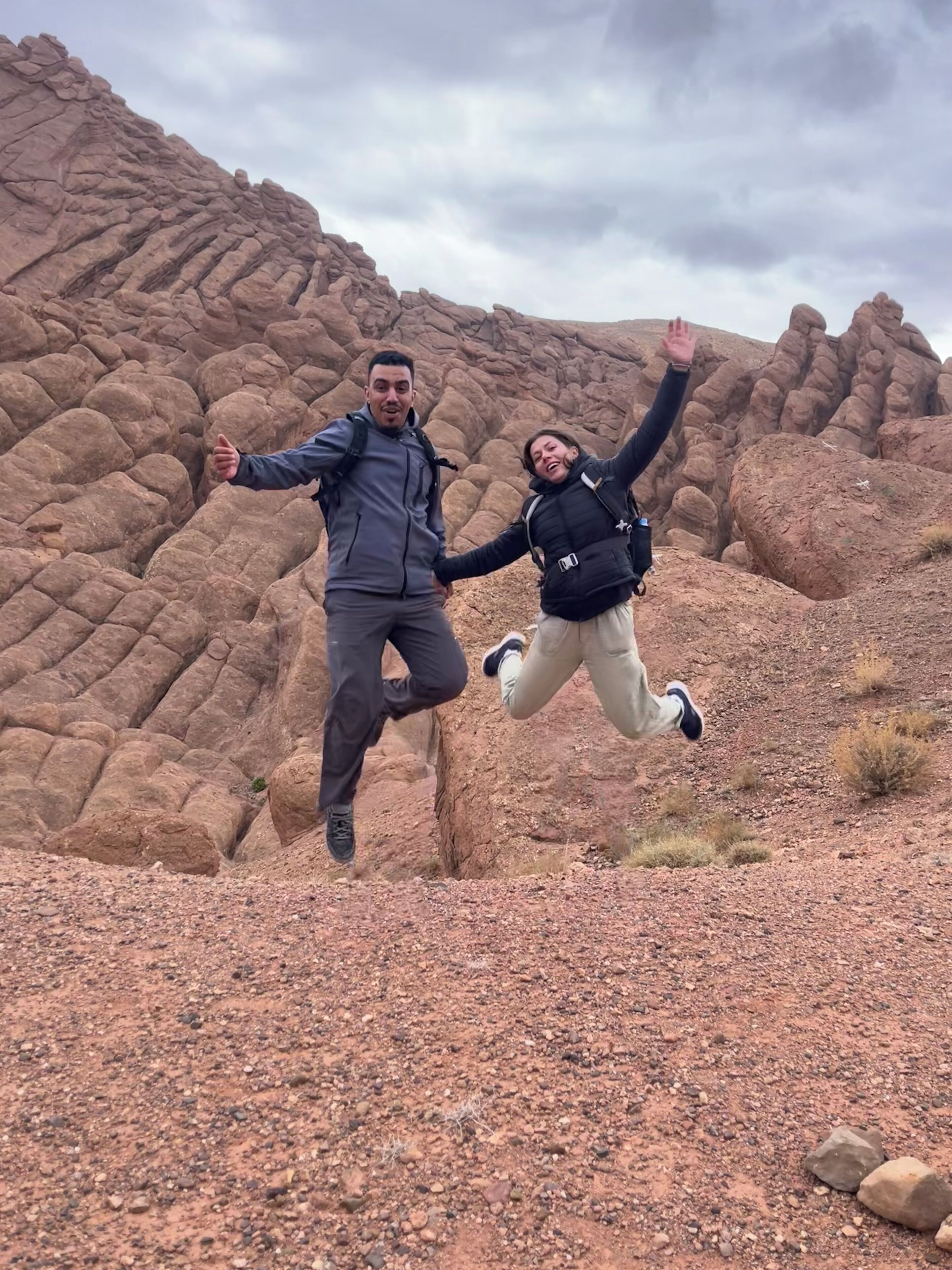 Dos personas saltando en un terreno rocoso al aire libre, con grandes rocas y un cielo nublado.