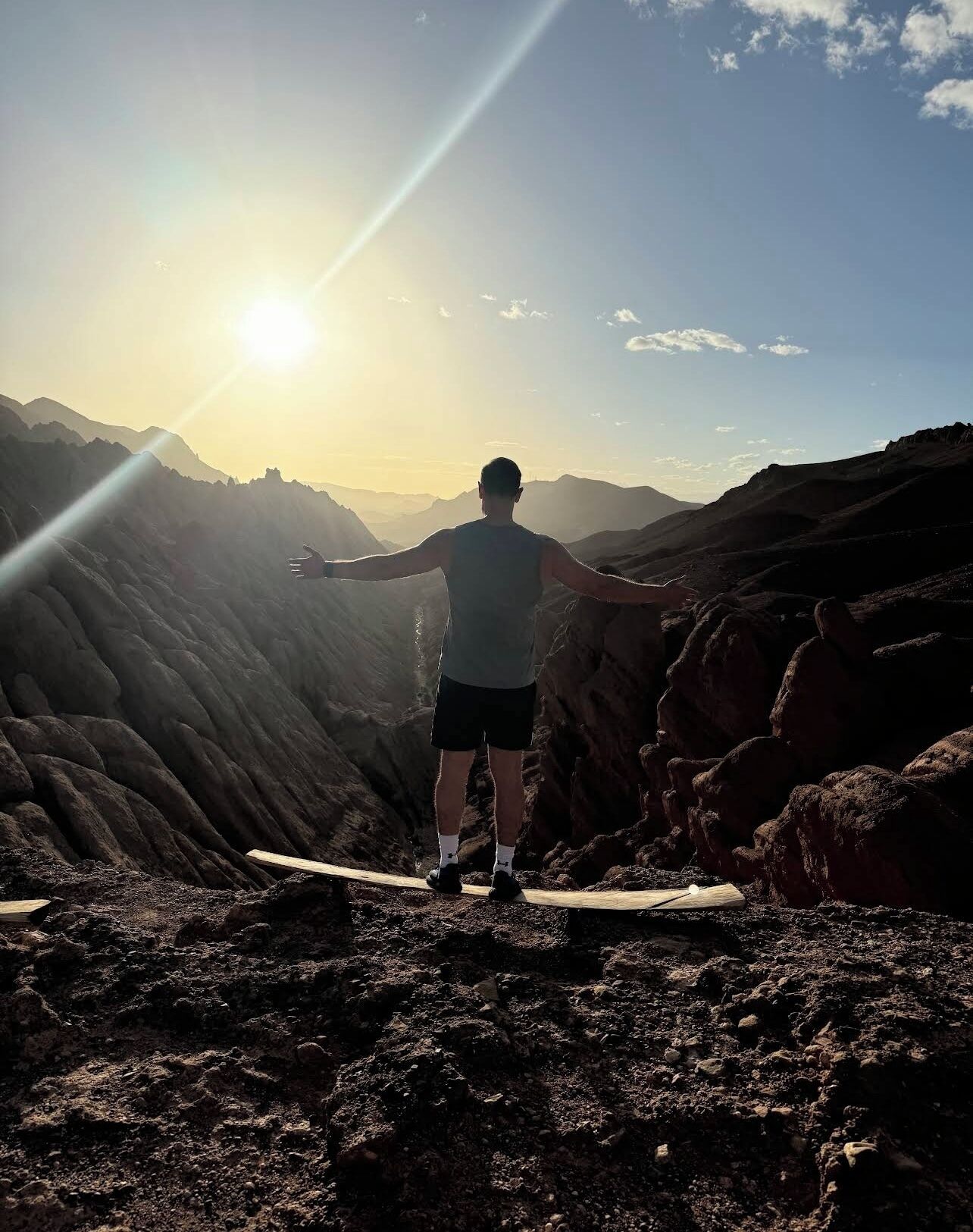 A person standing on a rocky outcrop with arms outstretched, overlooking a mountainous landscape during sunset.