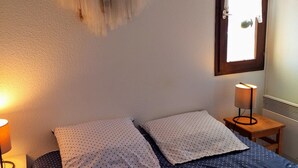 A bedroom with a bed, two pillows, a bedside lamp, a wall hanging, and a window with curtains. - Appartement vue sur mer (Soulac sur Mer)